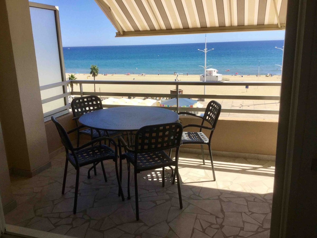 A spacious terrace is visible, featuring a round table surrounded by four chairs. The sea can be seen in the background, with gentle waves lapping at the shore. Awning provides shade to the seating area, enhancing comfort while enjoying the coastal view.