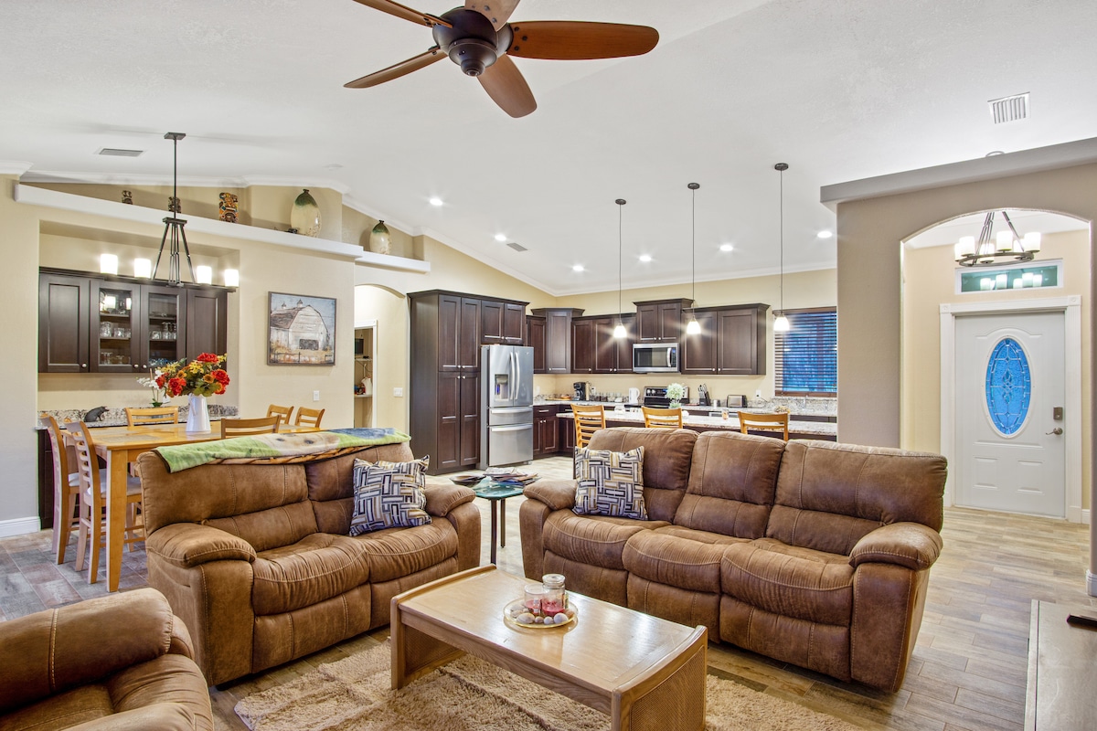 The spacious living area features plush seating arranged around a central coffee table. Light wood cabinetry contrasts with darker kitchen details in the background, while pendant lights illuminate the room. A soft rug adds warmth, and a decorative vase of flowers enhances the atmosphere.