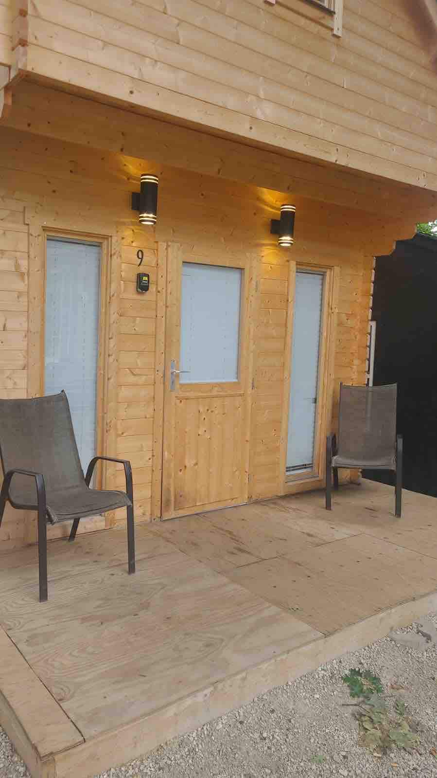 A wooden cabin facade features two main entry doors and is flanked by two simple chairs on a wooden deck. The exterior lighting is strategically placed above the doors, enhancing visibility at night.