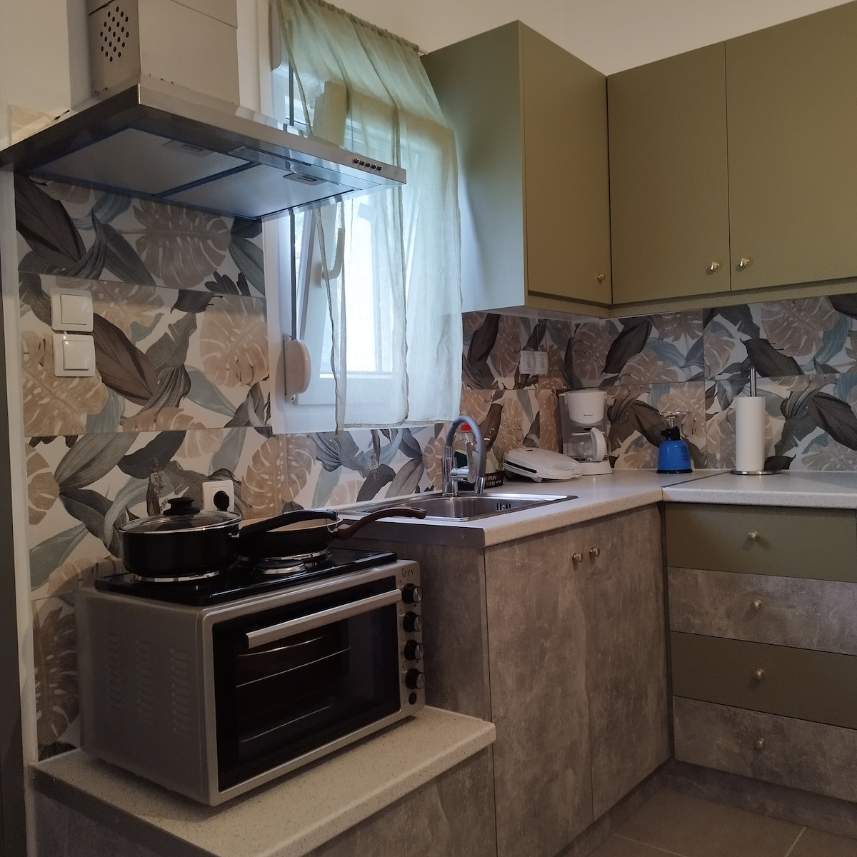 The kitchen features a blend of modern appliances and elegant cabinetry. A stovetop, oven, and sink are situated along a counter topped with a decorative backsplash. Natural light filters through a window adorned with a sheer curtain, enhancing the functional layout.