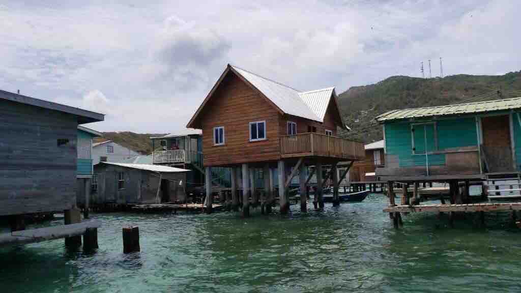 A wooden cabin stands elevated on stilts above the water, featuring a balcony and large windows. Surrounding structures exhibit a variety of colors and architectural styles, creating a charming coastal scene with a backdrop of green hills under a partly cloudy sky.