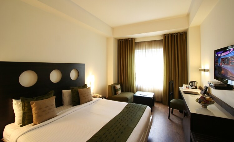 A well-appointed room features a spacious bed with an organized arrangement of decorative pillows. Natural light floods in through a large window, complemented by floor-length curtains. A small sitting area with a couch and table is visible, alongside a work desk equipped with a lamp and a TV.
