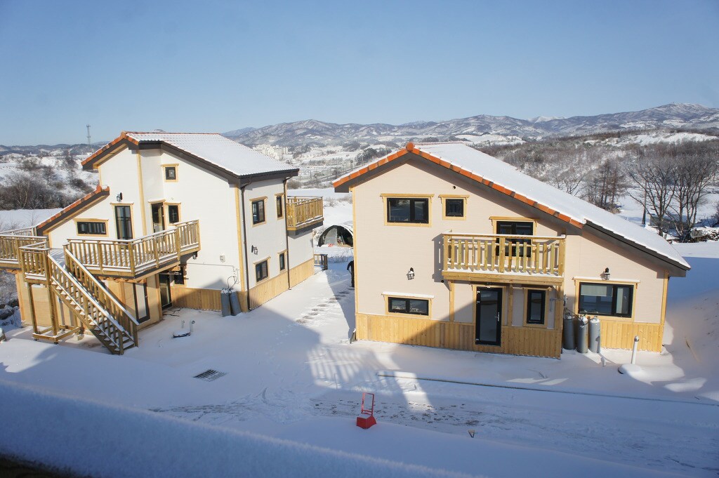 The image presents two newly constructed two-story log houses. Snow blankets the surrounding area, while the clear blue sky enhances the scene. Each house features wooden balconies and well-defined architectural lines, set against a backdrop of distant mountains.