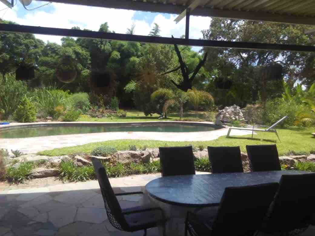 A serene outdoor area is visible, featuring a curvaceous swimming pool surrounded by lush greenery and varied plants. A patio dining table is situated in the foreground, accompanied by black chairs. The background showcases trees and a rock feature, enhancing the tranquil environment.
