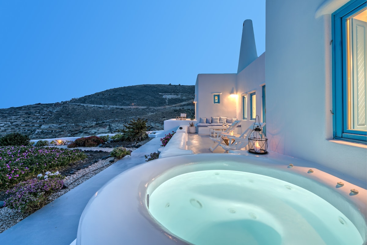 An outdoor area features a private built-in Jacuzzi, surrounded by tranquil landscaping with colorful flowers. Soft lighting and comfortable seating enhance the inviting atmosphere, while the serene hills in the distance provide a peaceful backdrop against a clear evening sky.