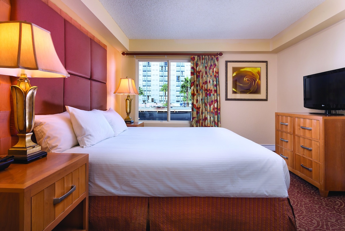 A well-appointed guest room features a spacious bed with crisp white linens. Nightstands with lamps flank the bed, and a sturdy wooden dresser is positioned nearby. A large window draped with colorful curtains allows natural light to enter, providing a view of the outside.