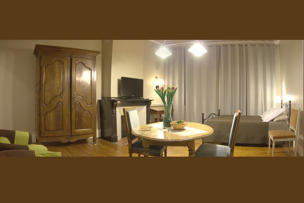 A cozy living area features a wooden wardrobe and a television mounted above a fireplace. A round dining table set for four is positioned beside the spacious bed, which is dressed in neutral tones. Soft light filters through floor-length curtains.