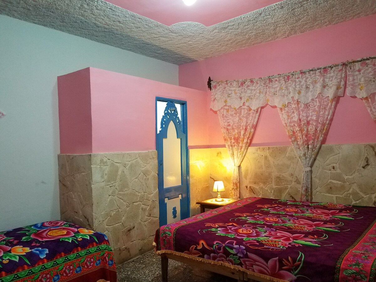 A sleeping area is characterized by colorful bedspreads featuring floral patterns. A spacious bed is complemented by a smaller bed nearby, both set against a backdrop of pink-painted walls. A small nightstand with a lamp provides soft illumination, while lace curtains frame a window.