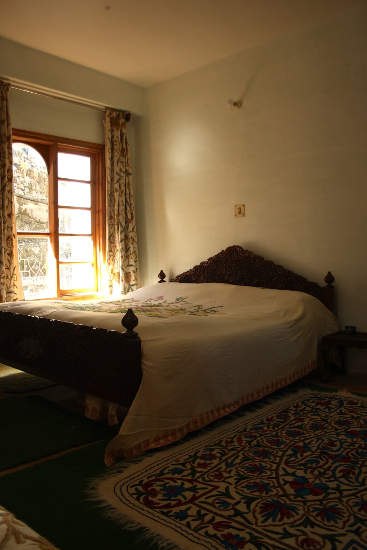 A spacious bedroom features a decorative wooden bed with a light-colored covering. Nearby, a large window allows natural light to fill the room, framed by patterned curtains. A colorful area rug adds warmth to the wooden flooring.