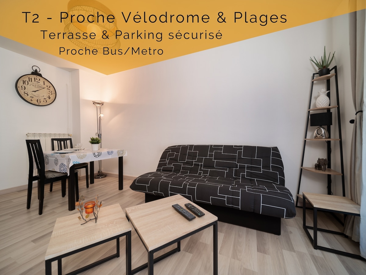 The living area features a dark patterned sofa bed, a table set for two, and a clock on the wall. Wooden side tables are situated near the sofa, providing a balanced and functional arrangement. Natural light illuminates the space through a window.