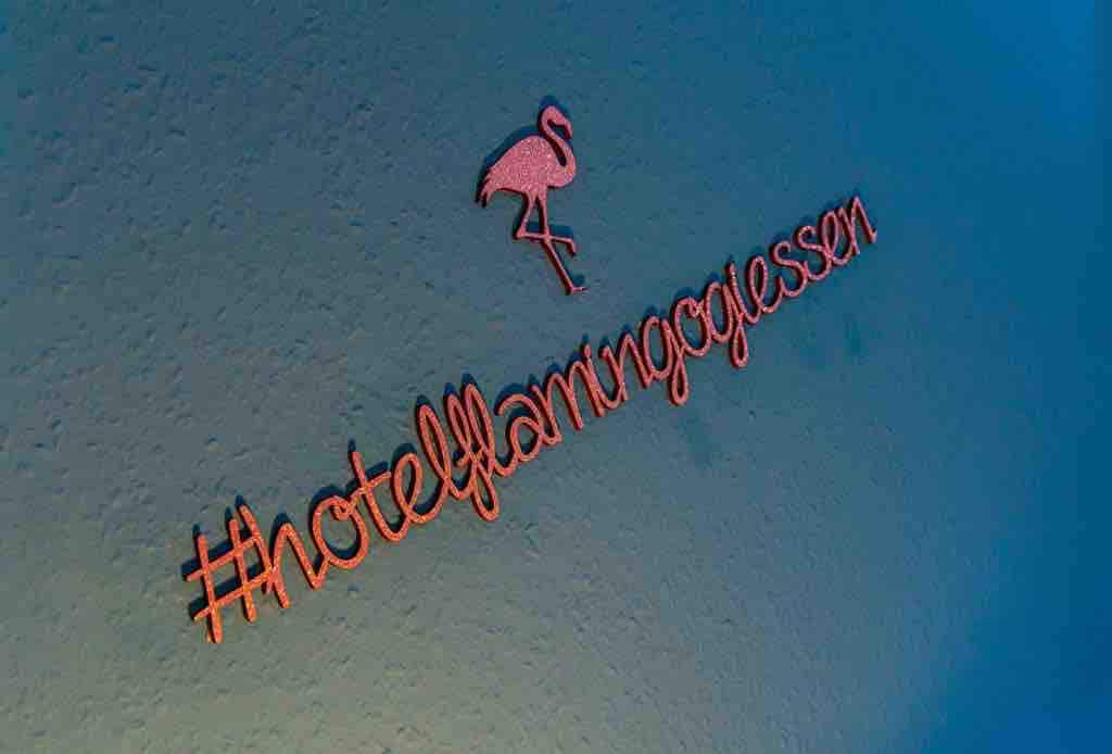 A decorative wall displays a hashtag with the words 'hotel flamingo giessen' in a playful font. A pink flamingo silhouette complements the vibrant orange lettering, creating a lively atmosphere.