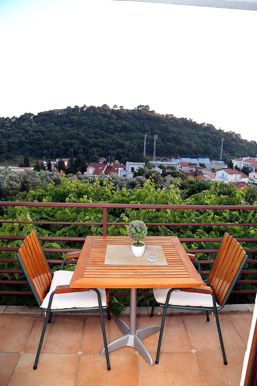 An outdoor balcony features a wooden table set with two chairs, overlooking lush greenery and a view of the nearby town. A small potted plant decorates the table, adding a touch of nature to the serene atmosphere.