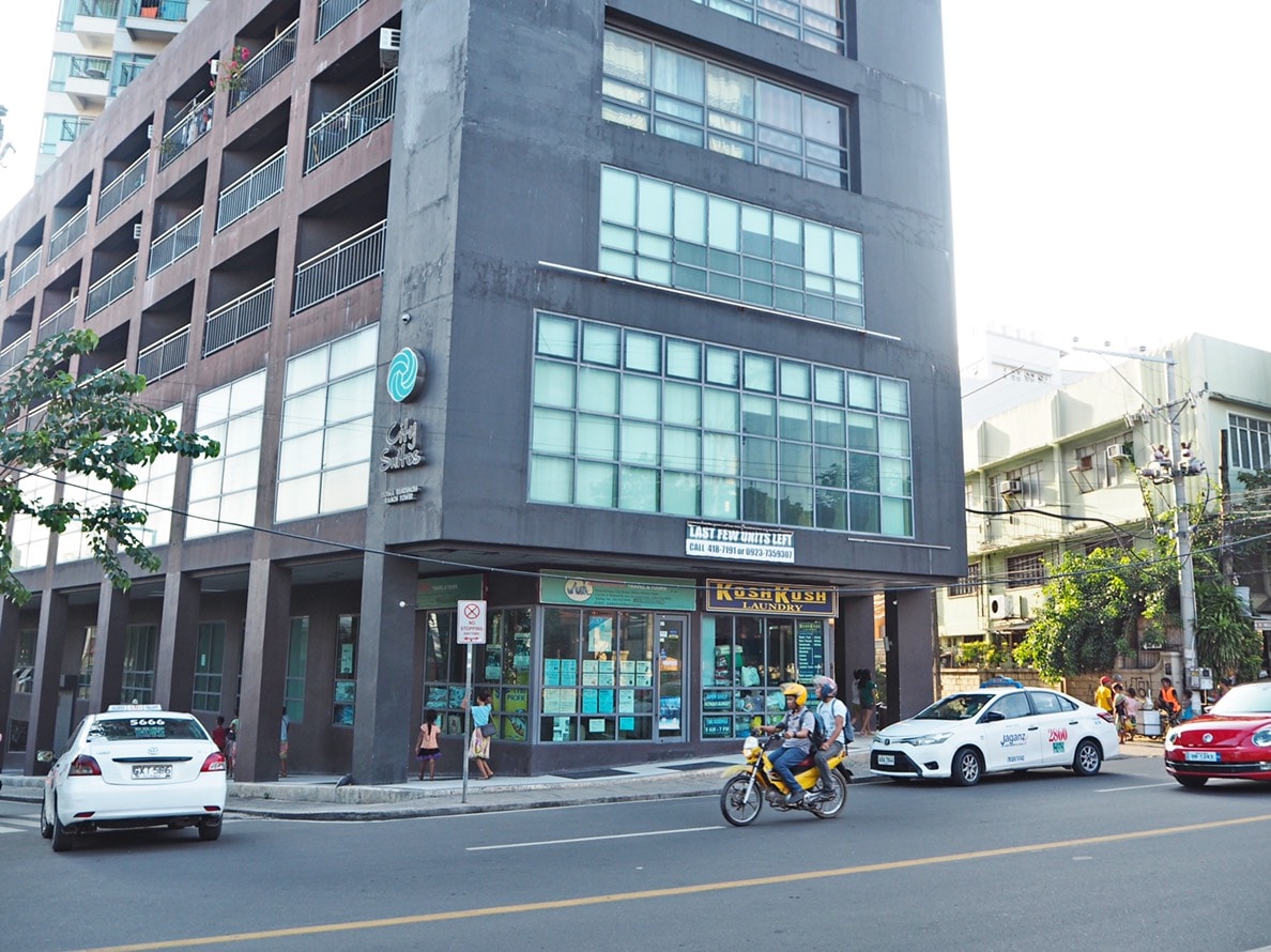 The exterior of a modern building is displayed, showcasing large glass windows on the upper floors. Shops on the ground floor are visible, along with parked cars and a motorcycle passing by on the street, contributing to the urban atmosphere of the area.