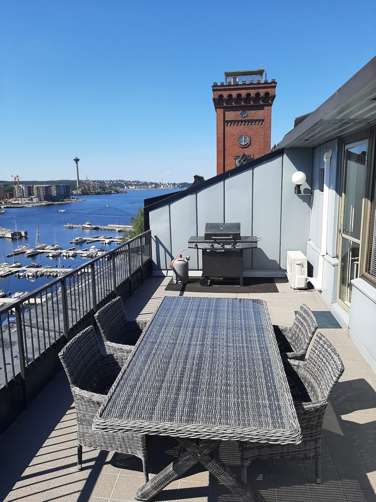 A spacious terrace features a large outdoor dining table surrounded by six woven chairs. A grill is positioned nearby, offering scenic views of the waterfront and the city skyline, including a prominent clock tower in the background.