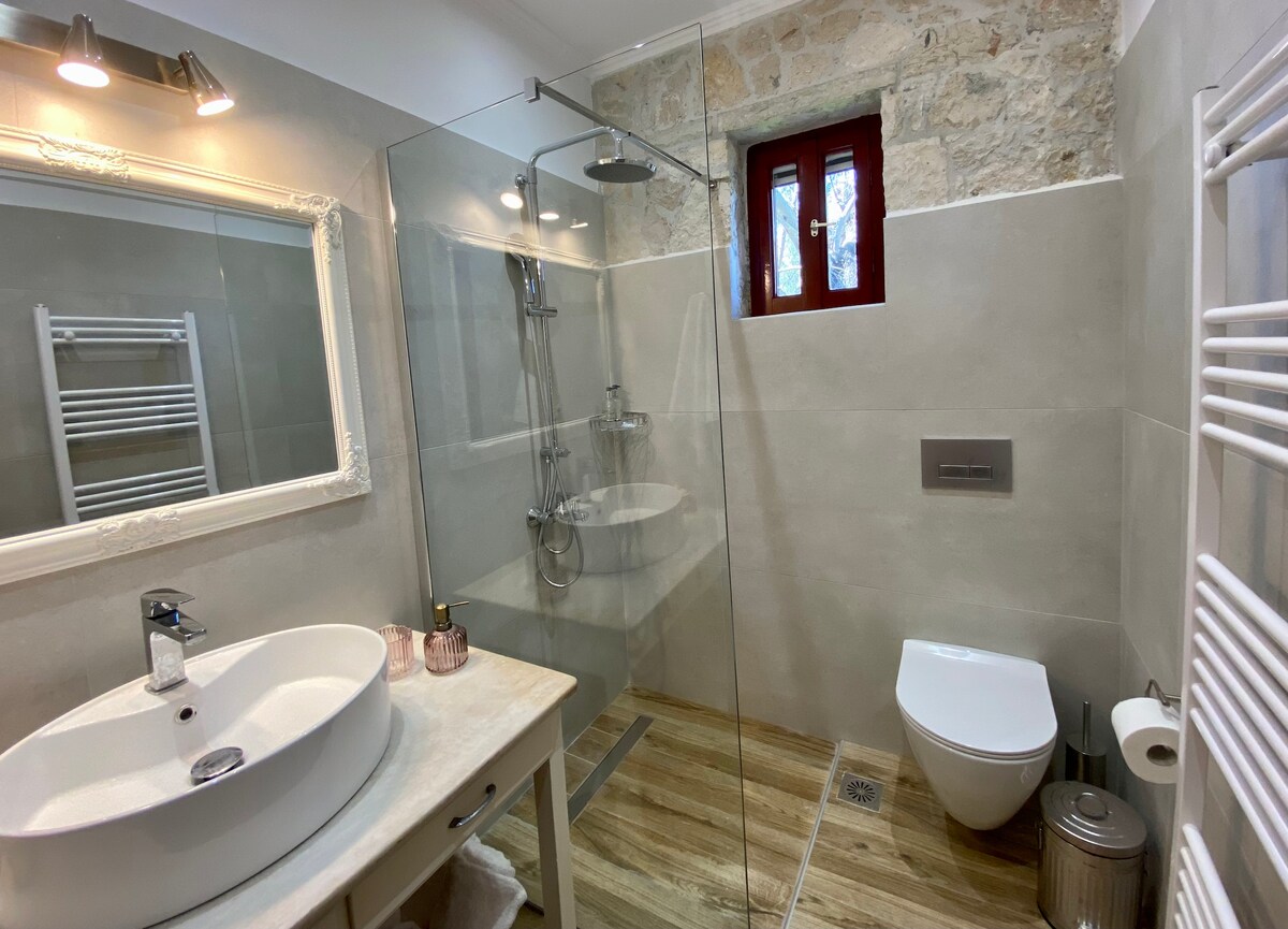 A modern bathroom features a glass-enclosed shower and a sleek toilet. A round sink is positioned on a wooden vanity, with a wall-mounted mirror above. Natural light filters through a small window, highlighting the stone wall and wood-effect flooring for a clean, inviting space.
