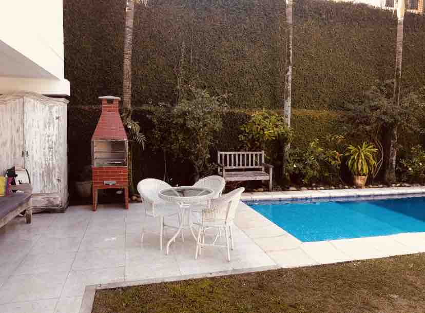 A serene outdoor space features a swimming pool complemented by a well-maintained garden. A barbecue grill is positioned beside a wooden bench, and a small round table with four chairs invites relaxation. Lush greenery serves as a backdrop, enhancing the tranquil atmosphere.