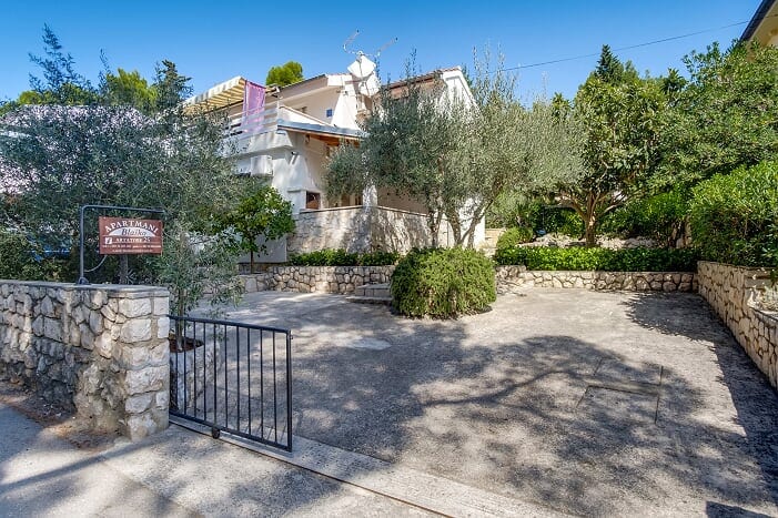 An inviting entrance to the property is framed by lush olive trees and shrubs. A spacious outdoor area is paved with stone, featuring a gated entry that enhances privacy. A clear blue sky complements the tranquil surroundings, creating a peaceful first impression.