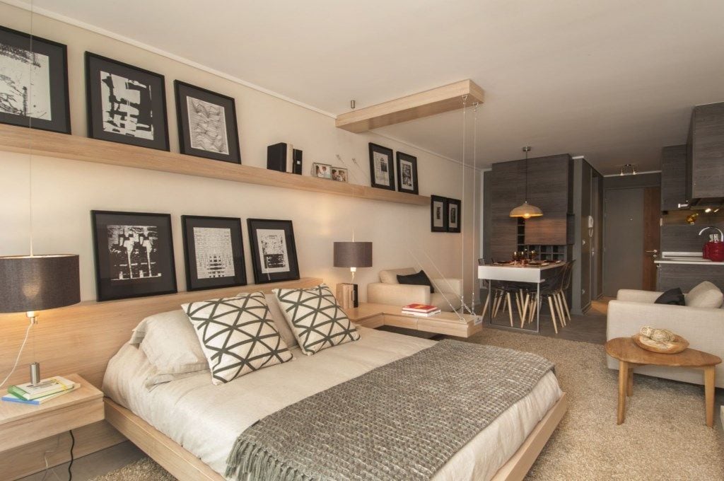 A modern studio room features a spacious bed with soft bedding and patterned pillows. Wall-mounted shelves display framed artwork, while a cozy seating area is arranged nearby. The open kitchen is visible, equipped with contemporary appliances and a dining space with stylish chairs.
