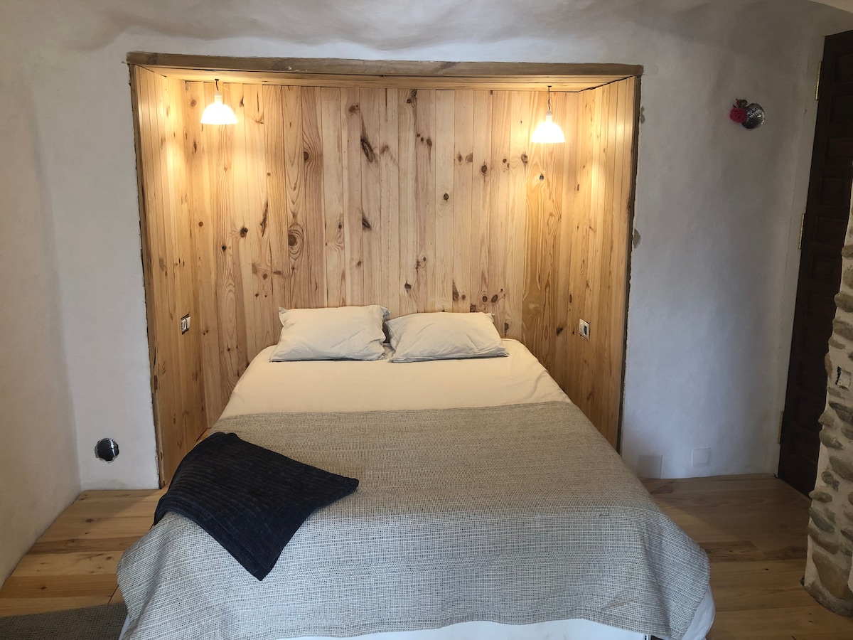 A cozy sleeping area features a wooden accent wall with two softly glowing wall lights. A spacious bed is topped with neutral linens and decorative pillows, while a textured throw blanket adds a touch of warmth. Natural materials enhance the cave-like ambiance.