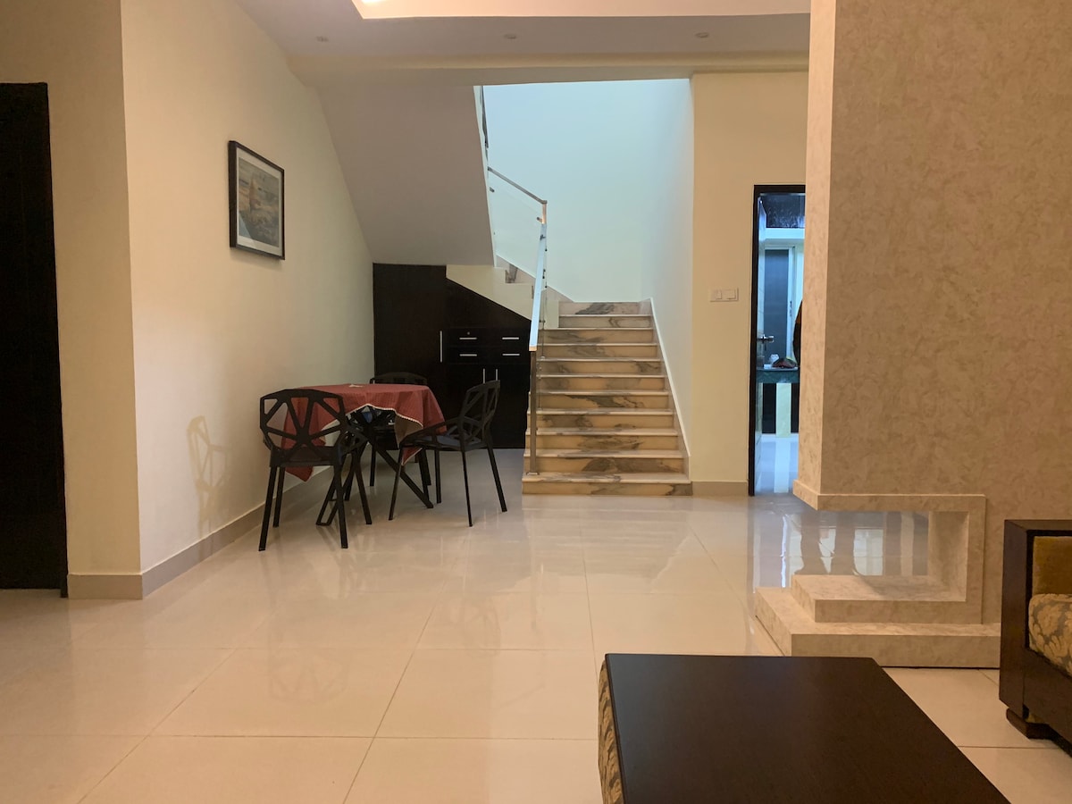 A spacious living area is seen, featuring a dining table with four chairs. A staircase, framed by a sleek railing, leads to an upper level. The floor is tiled, reflecting natural light that fills the room. Neutral walls complement the minimalist decor.
