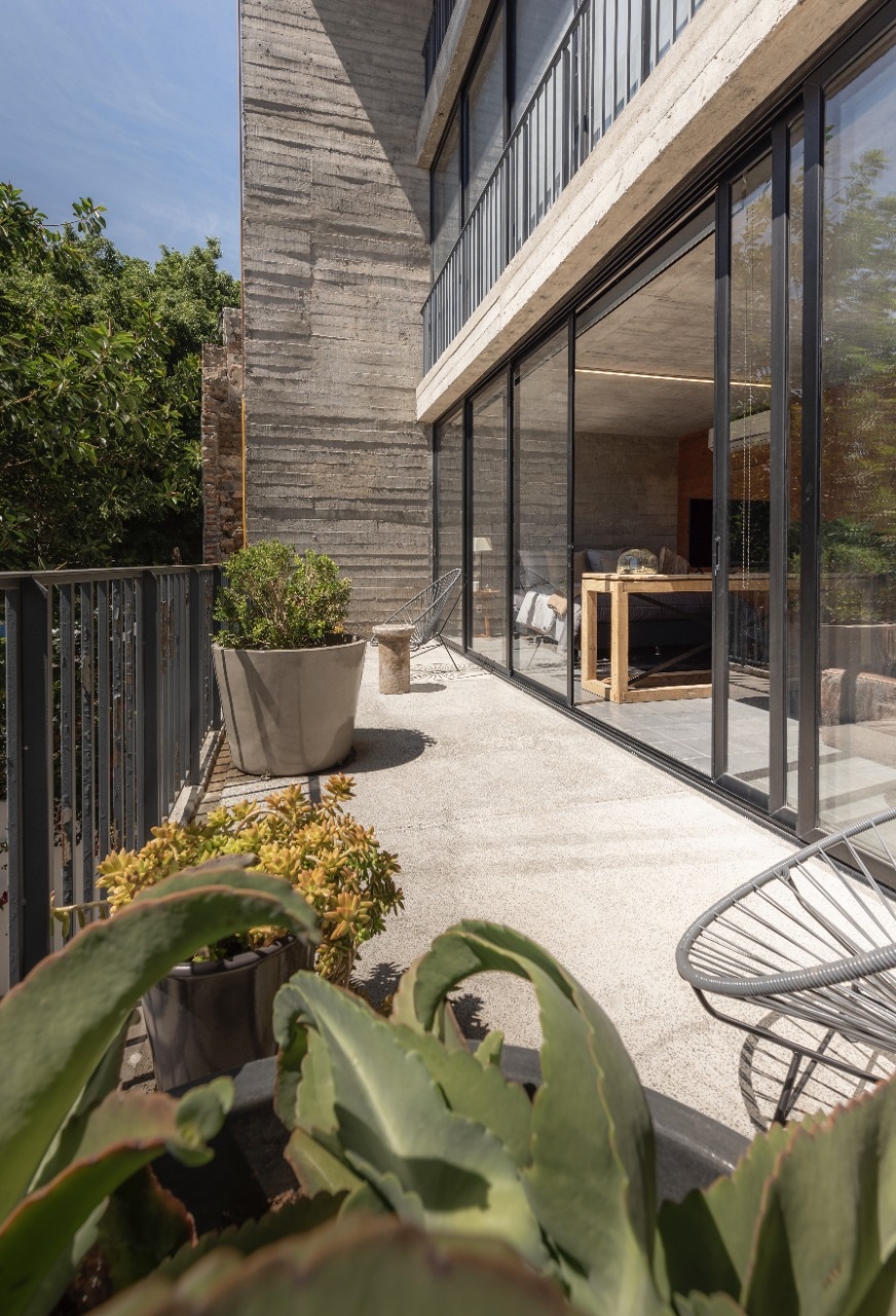 A spacious balcony is depicted, featuring a clean concrete floor and complemented by decorative planters with green foliage. Large glass doors offer views into the interior, while sleek outdoor chairs are positioned for relaxation. Lush greenery surrounds the area, enhancing the inviting atmosphere.