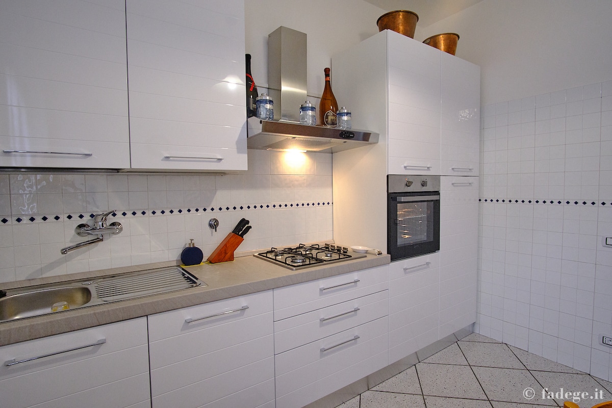 A modern kitchen space is equipped with sleek white cabinets and a countertop. Stainless steel appliances include an oven and a gas cooktop. Kitchen utensils are arranged neatly, and a sink is located adjacent to the cooking area, with decorative elements enhancing the overall functionality.