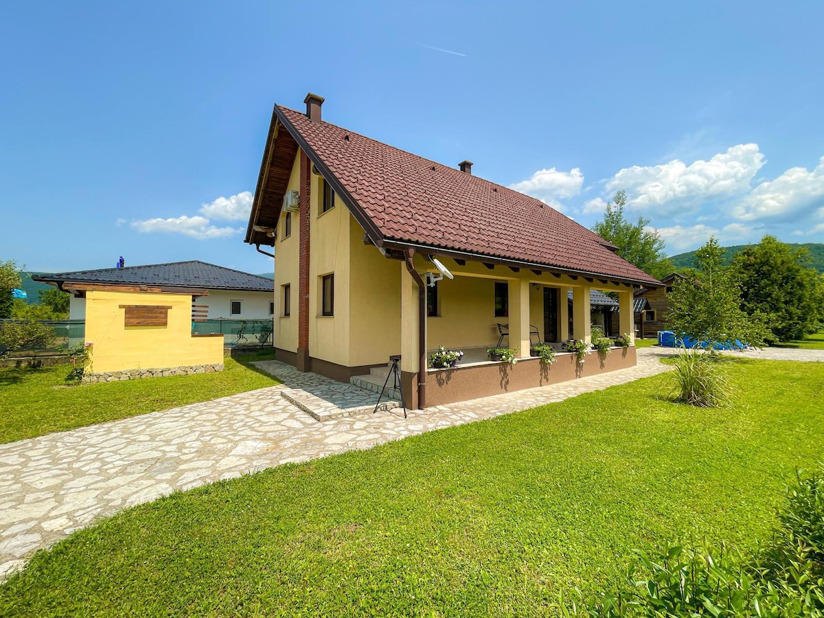 A two-story vacation home features a brown tile roof and a warm yellow exterior. A stone pathway leads to a covered porch with flowerboxes. Surrounding greenery and a blue sky filled with clouds create a peaceful outdoor ambiance.