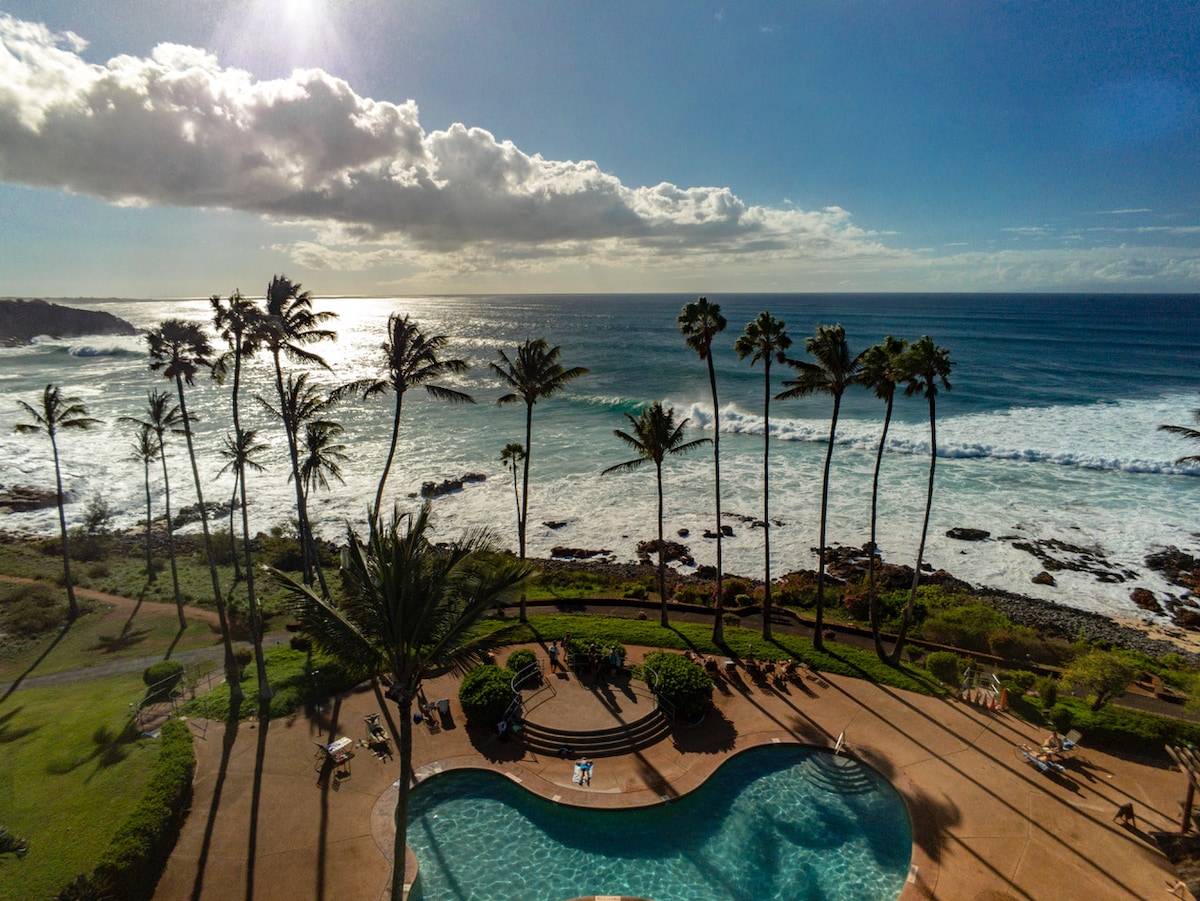 An oceanfront pool is surrounded by palm trees, providing a scenic view of the beach and waves. The bright blue water of the pool reflects the clear sky, while the gentle surf adds a tranquil sound to the scene.