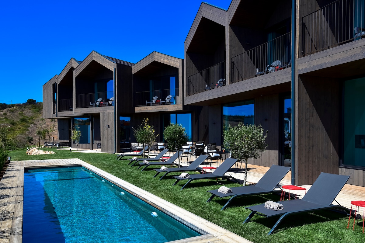 A sleek outdoor area showcases a turquoise swimming pool bordered by well-maintained grass. Lounge chairs are arranged around the pool, providing options for relaxation. The modern structure features multiple levels, with large windows and balconies allowing for ample natural light and views of the surrounding landscape.