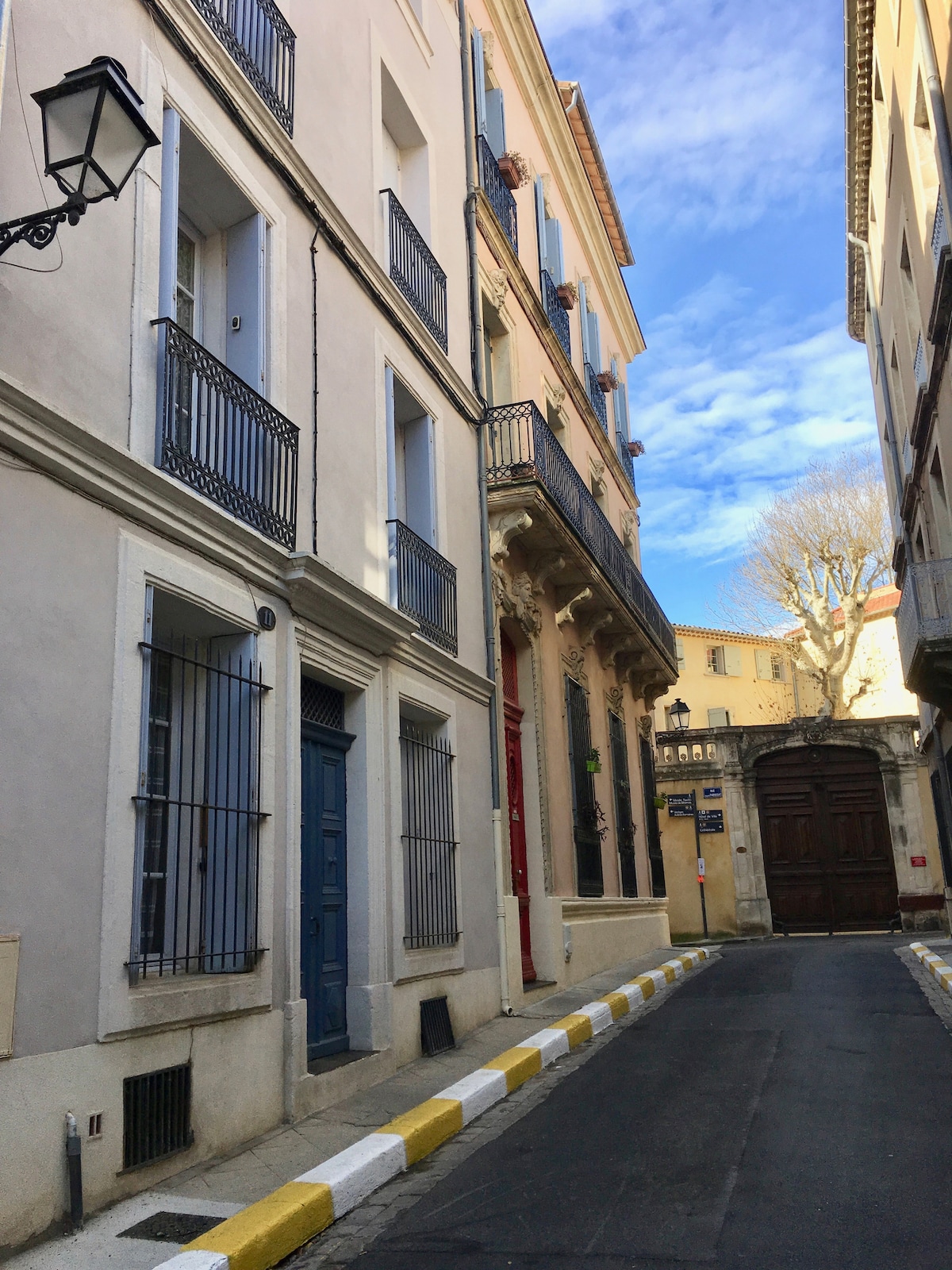A narrow street is flanked by charming buildings featuring balconies and decorative elements. The sky is bright with scattered clouds, casting soft light over the scene. A rustic wooden door stands at the end of the lane, inviting exploration of the surrounding area.
