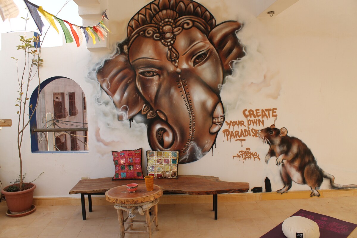 A vibrant communal space is adorned with a large mural, showcasing artistic expressions. A wooden bench set against the wall is accompanied by colorful cushions and a small decorative table. Natural light seeps through an arched window, enhancing the welcoming atmosphere.