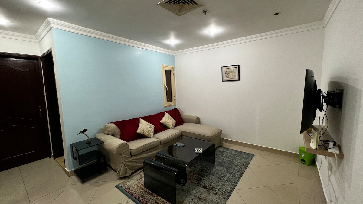 A modern living room is presented with a light blue accent wall, featuring a comfortable sofa in neutral tones with decorative pillows. A dark coffee table is positioned in the center, while a flat-screen TV is mounted on the wall nearby. Natural light enters through a window.