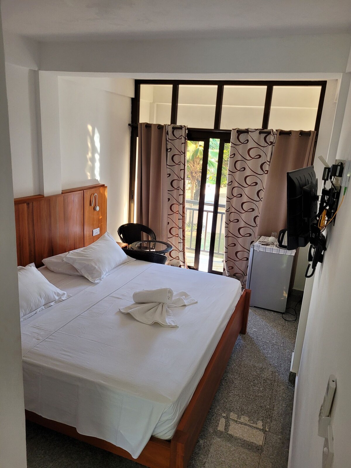 A brightly lit bedroom features a spacious bed with white linens and two pillows. Natural light filters in through large windows, adorned with light curtains. A small table and chair are positioned near the window, with a television mounted on the wall and a mini-fridge visible.