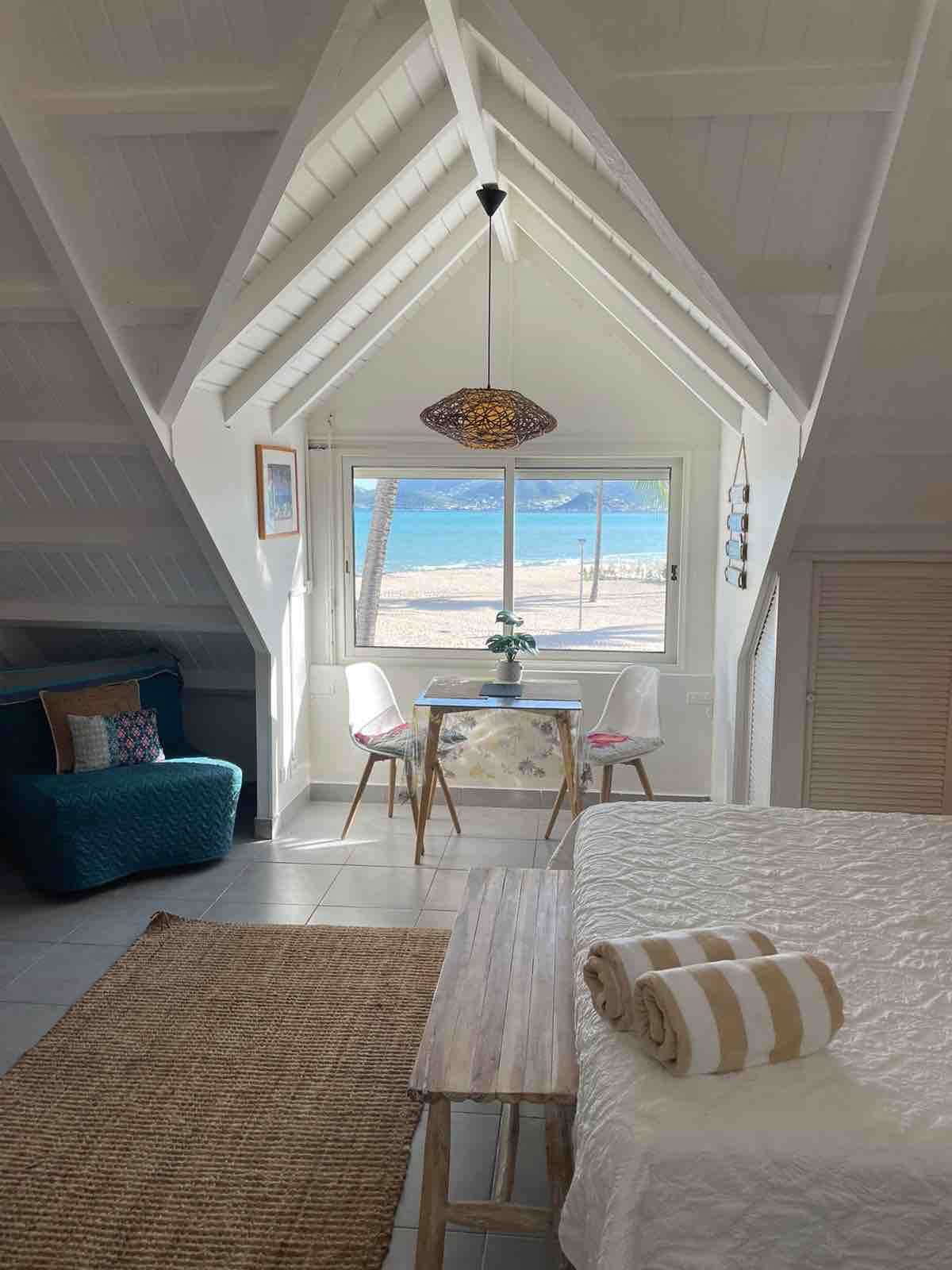 A bright studio space features vaulted ceilings and a window showcasing views of the Caribbean Sea. A small dining table is accompanied by clear chairs, while a cozy seating area is arranged nearby. Soft linens are neatly placed on the bed, complemented by decorative towels.