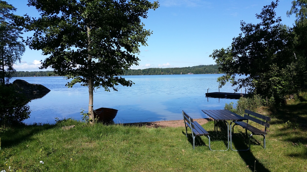 A tranquil lakeside scene features a grassy area with a picnic table and benches. Nearby, a small rowboat is visible, along with a wooden dock extending into the calm water. Lush trees frame the view, creating a serene atmosphere by the lake.