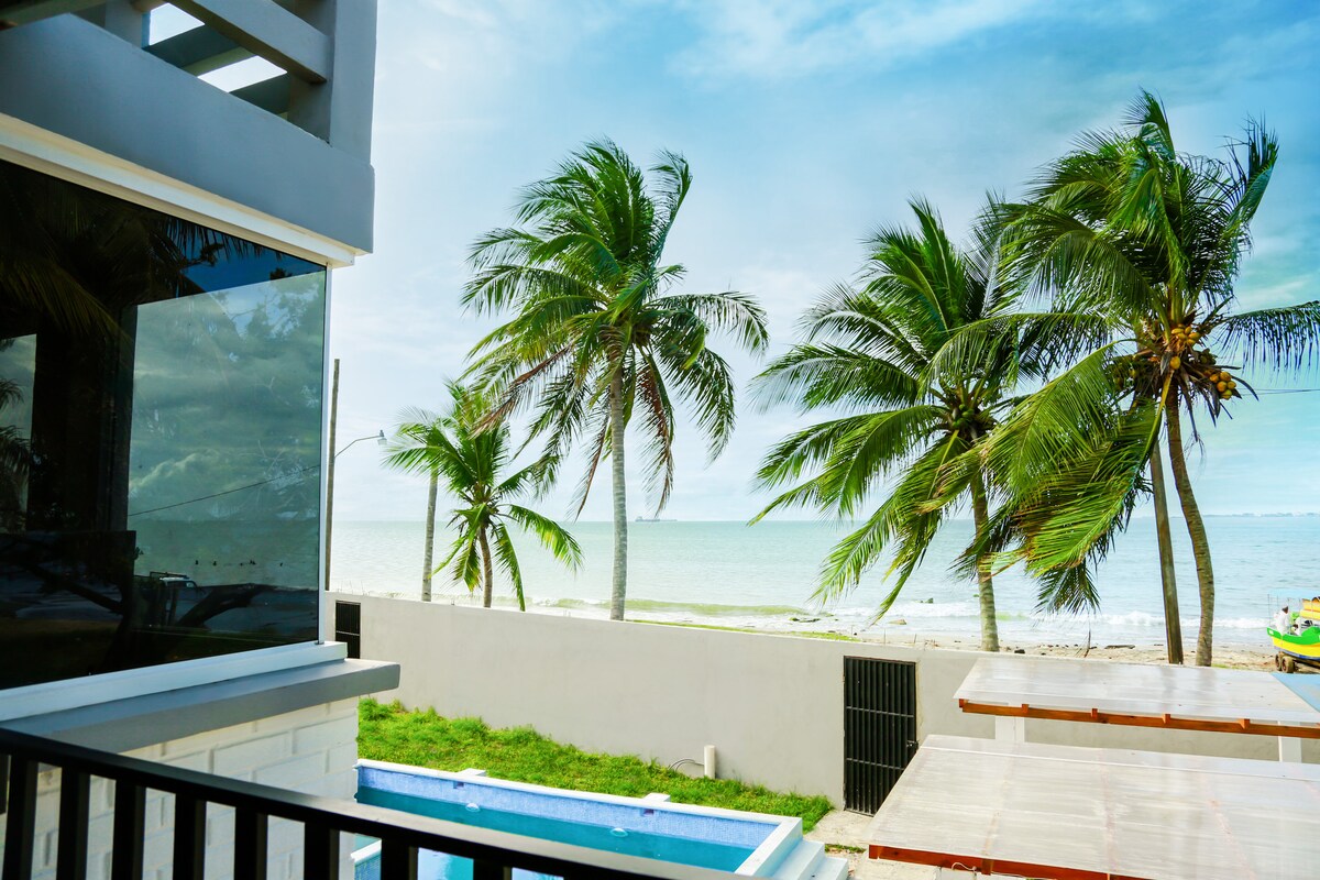 A clear view of the ocean can be seen from the property, framed by swaying palm trees. The image features a tranquil pool beside a grass area, with the sandy beach visible in the distance, creating a serene coastal atmosphere.
