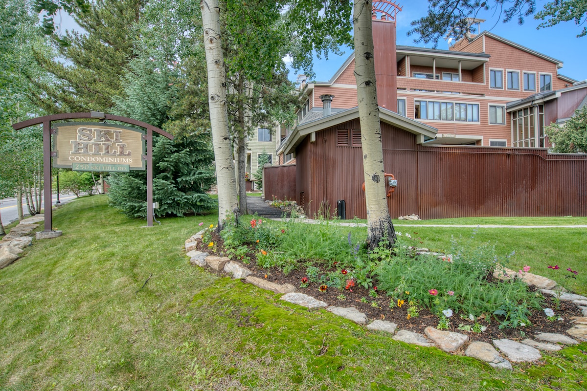 The entrance to the Sea Hill Condominiums is showcased, featuring well-maintained landscaping with a variety of flowers and stones. The three-story building, constructed with warm wood and stone accents, is surrounded by scenic greenery and tall trees, creating a welcoming environment.