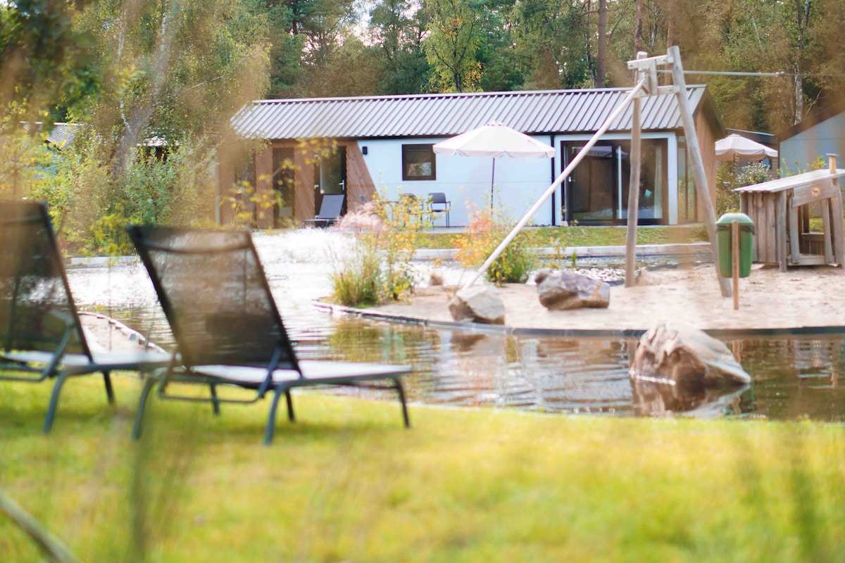 A modern chalet is set within a natural landscape, featuring large windows and a sloped roof. A small pond is positioned nearby, with rocks and sandy areas. Two lounge chairs are arranged on grass, offering a view of the play area, which includes a swing set.