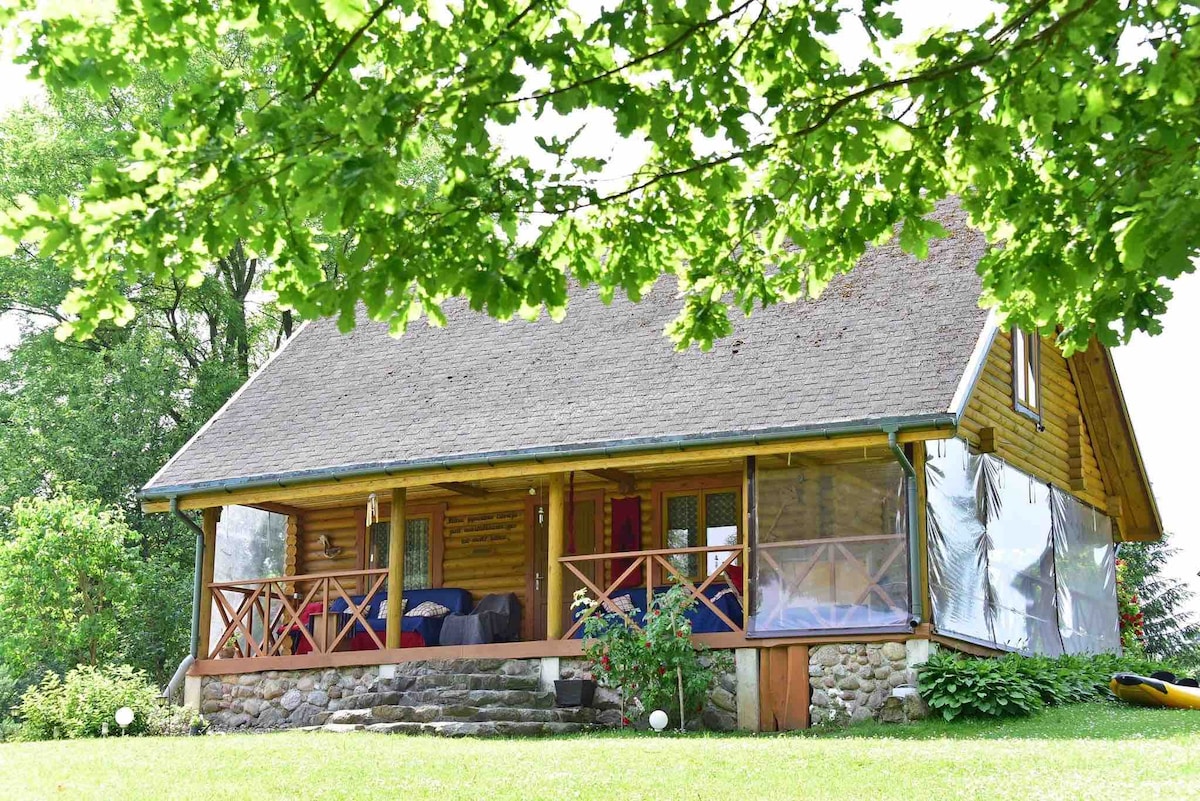 A charming wooden cottage is nestled among greenery, featuring a welcoming front porch with wooden railings. The structure showcases large windows on either side, allowing for ample natural light. A stone pathway leads to the entrance, while vibrant foliage provides a serene setting.