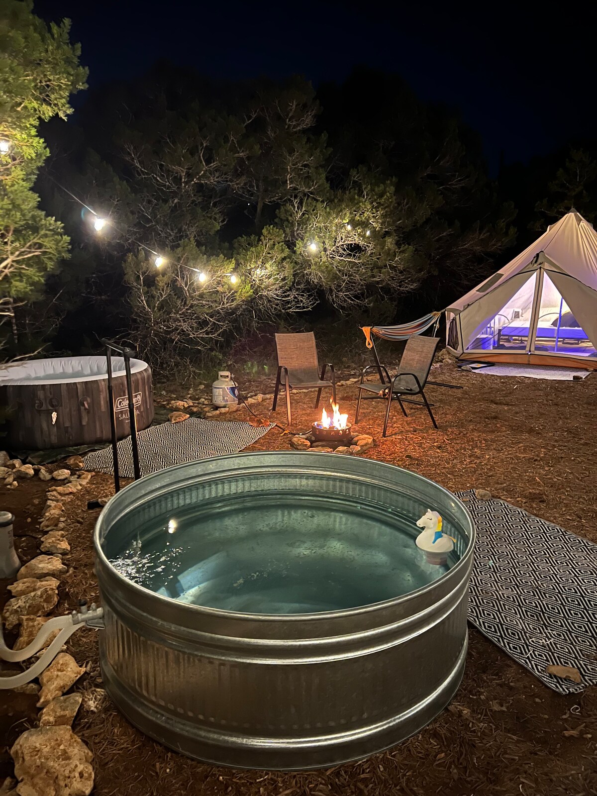 A cozy outdoor setting is presented in the evening, featuring a stock tank dipping pool and a private hot tub. Several seating options including two chairs and a hammock surround a small fire pit, with ambient string lights enhancing the inviting atmosphere.