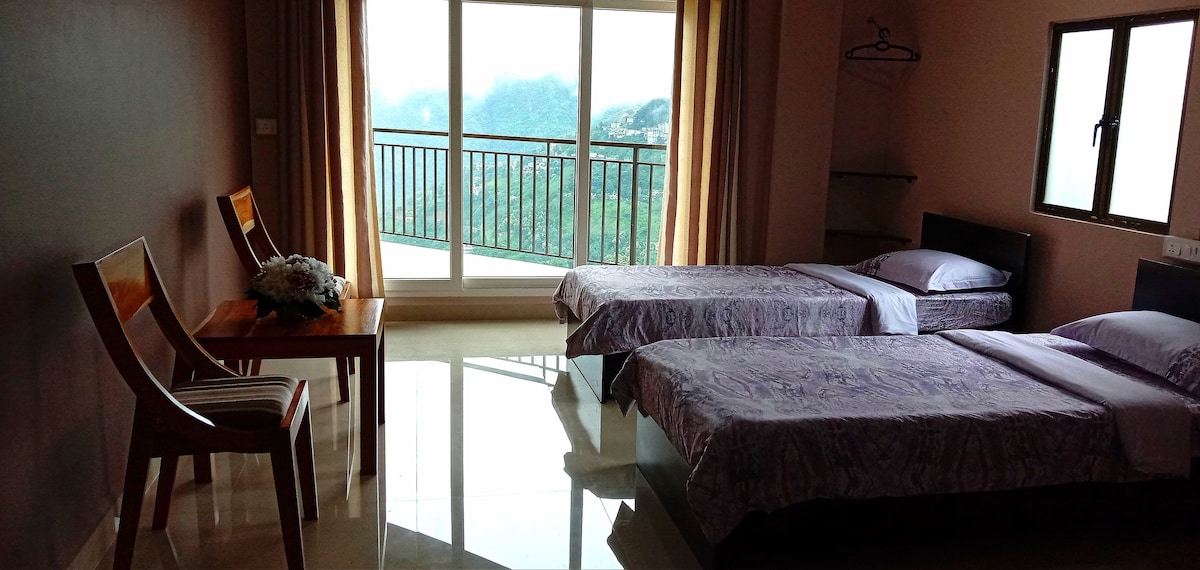 A spacious guest room features two single beds with lavender bedding, positioned beside a small wooden table and a chair. Large glass doors provide a view of the surrounding landscape, with natural light brightening the room. Soft curtains frame the window.