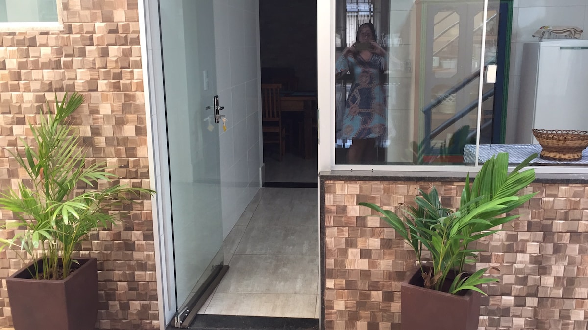 An entrance is framed by two large potted plants, enhancing the welcoming feel of the space. A glass door allows visibility into the interior, while a textured brown wall provides a contrasting backdrop. The floor is tiled and features a subtle pattern leading indoors.