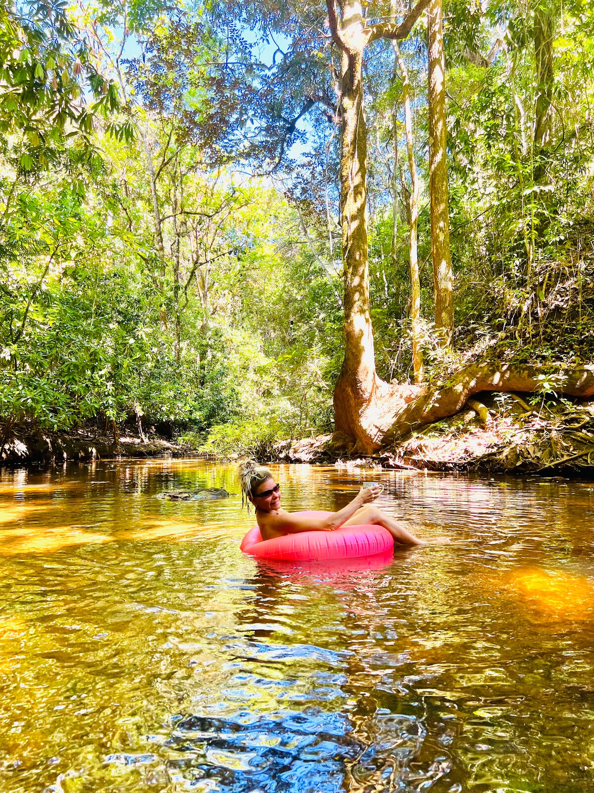 A serene natural setting is depicted, featuring a clear river winding through lush greenery. Sunlight filters through the canopy, creating dappled reflections on the water's surface. A red inflatable tube floats gently, inviting relaxation amidst the tranquil surroundings of a tropical jungle.