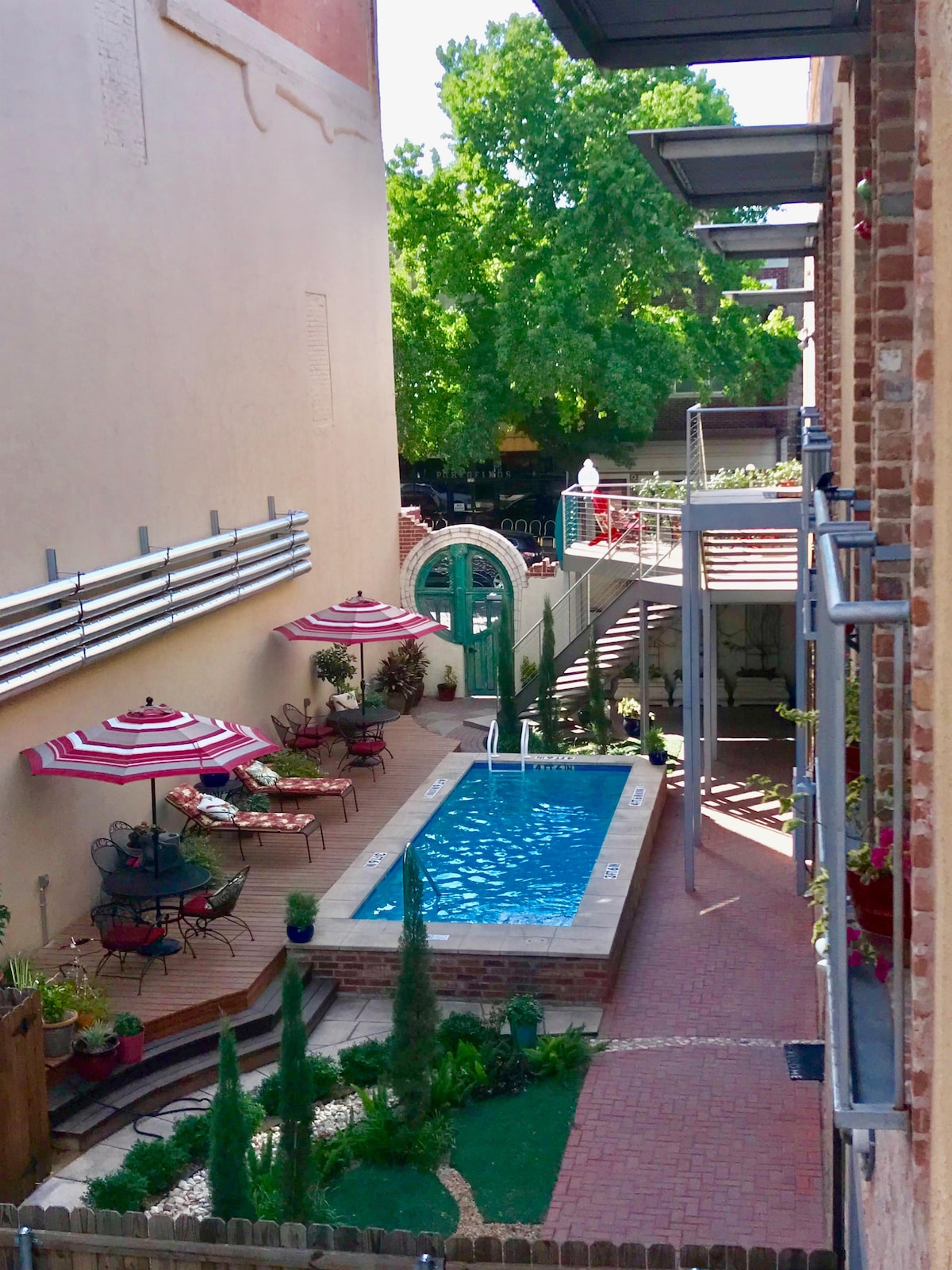 A heated courtyard pool is framed by lounge chairs and small tables, surrounded by landscaped greenery. Two striped umbrellas provide shade for the seating area. The view captures the serene atmosphere between the loft units and the pool, highlighting the inviting outdoor space.