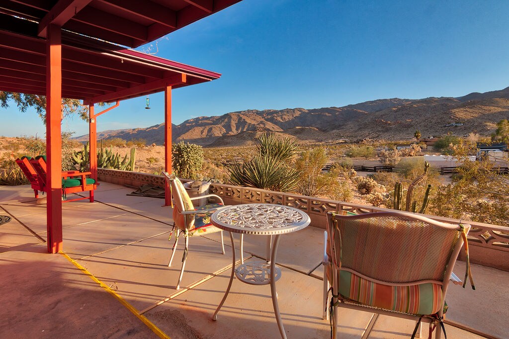 A patio area features colorful chairs alongside a small table, set against a backdrop of rolling desert hills. The scene is illuminated by warm sunlight, with distant mountains and cacti visible in the landscape.