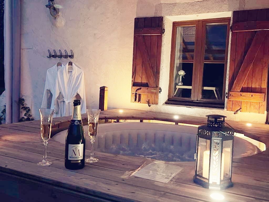 An outdoor spa area is displayed, featuring a cozy hot tub surrounded by wooden decking. A bottle of champagne is positioned beside two champagne flutes on the table. Soft lighting from the lantern and window creates a warm ambiance.