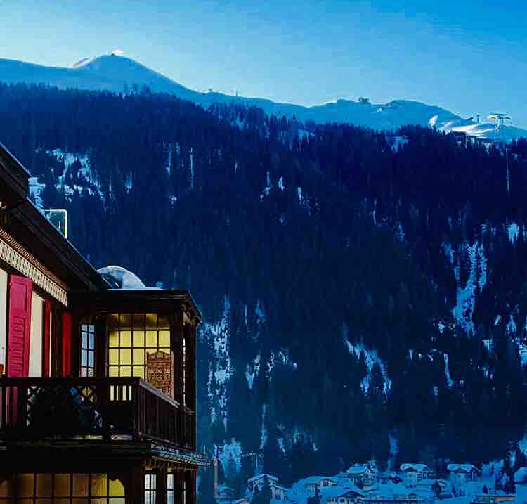 A stunning view captures the snowy mountains in the background, with trees blanketed in white. The building's balcony reveals wooden accents, and several large windows reflect the morning light. The serene landscape offers a glimpse of the surrounding winter beauty.