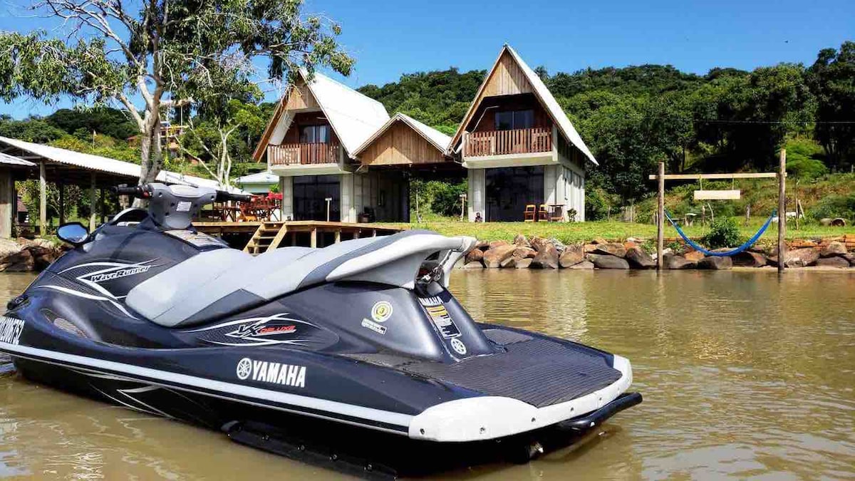 A jet ski rests in the calm water, with two spacious cabins visible in the background. The cabins feature wooden balconies and are surrounded by greenery, while a hammock is seen hanging nearby, inviting relaxation by the water's edge.