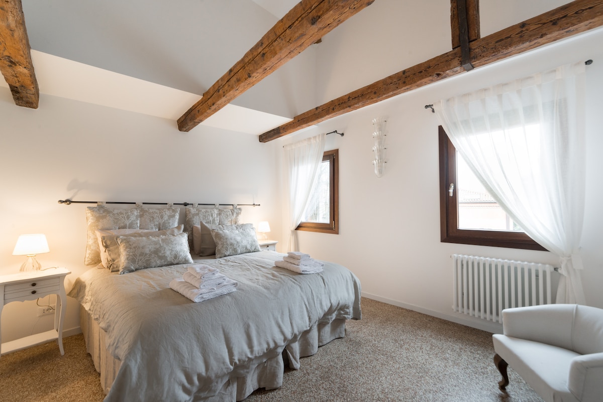 A spacious bedroom features two beds dressed in light, neutral linens, flanked by bedside tables with soft lighting. Natural light filters through sheer curtains, illuminating the wooden beams above. A comfortable chair sits in the corner, complementing the room's inviting atmosphere.