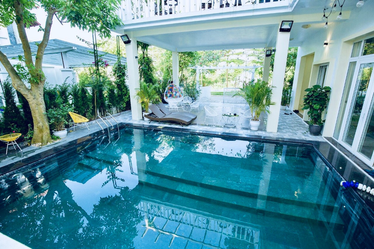 An inviting pool area is surrounded by lush greenery and sleek stone flooring. Sun loungers are positioned beside the pool, reflecting the light from the overhead fixtures. Large windows and a balcony provide an unobstructed view of the tranquil outdoor space.